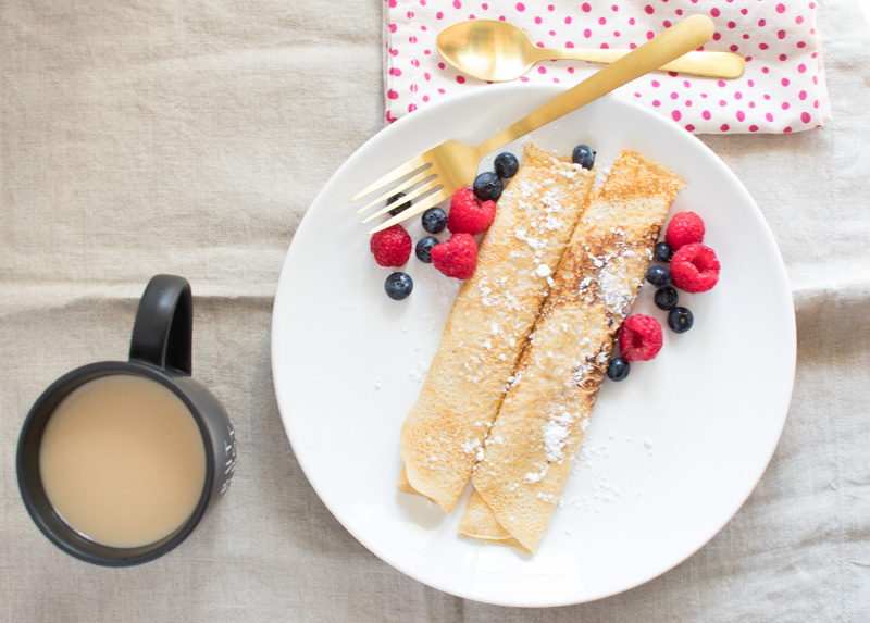 Overhead view of two crepes on a plate with fruit and a cup of coffee