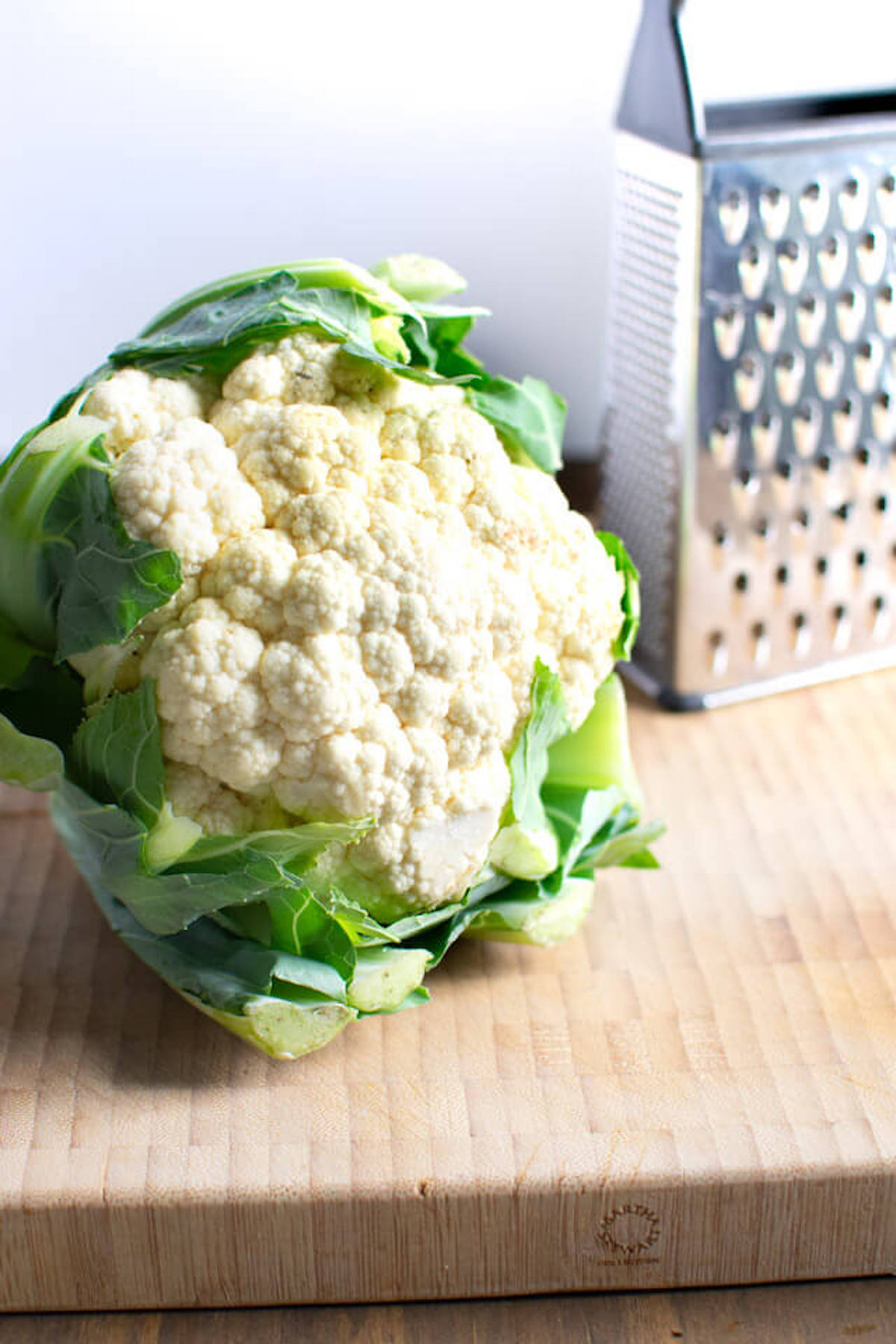 Head of cauliflower on wooden cutting board with box grater behind it