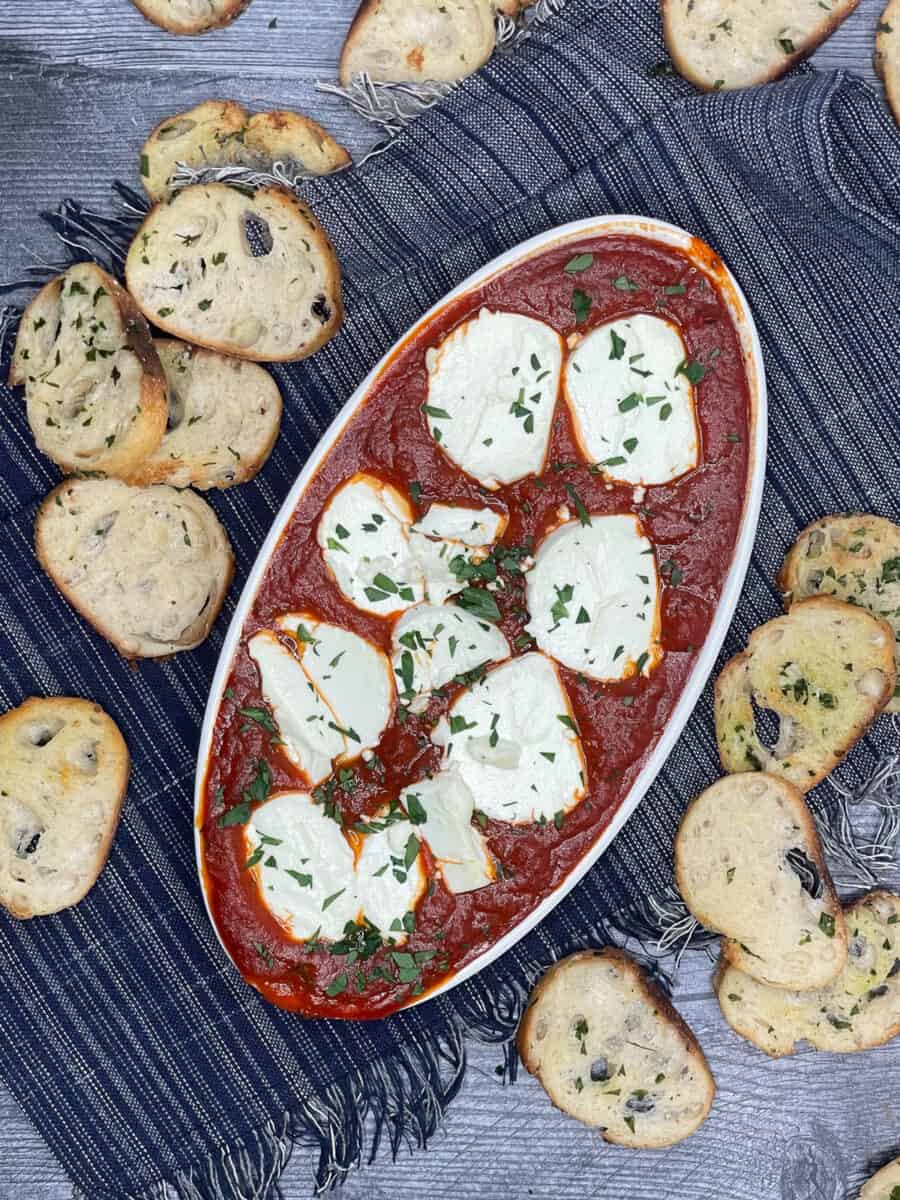 overhead view of baked goat cheese dip in a white oval dish with crostini next to it