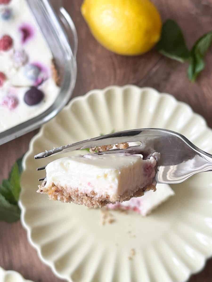 Creamy berry cheesecake on a fork, served on a scalloped plate, with fresh lemon and berries in the background.