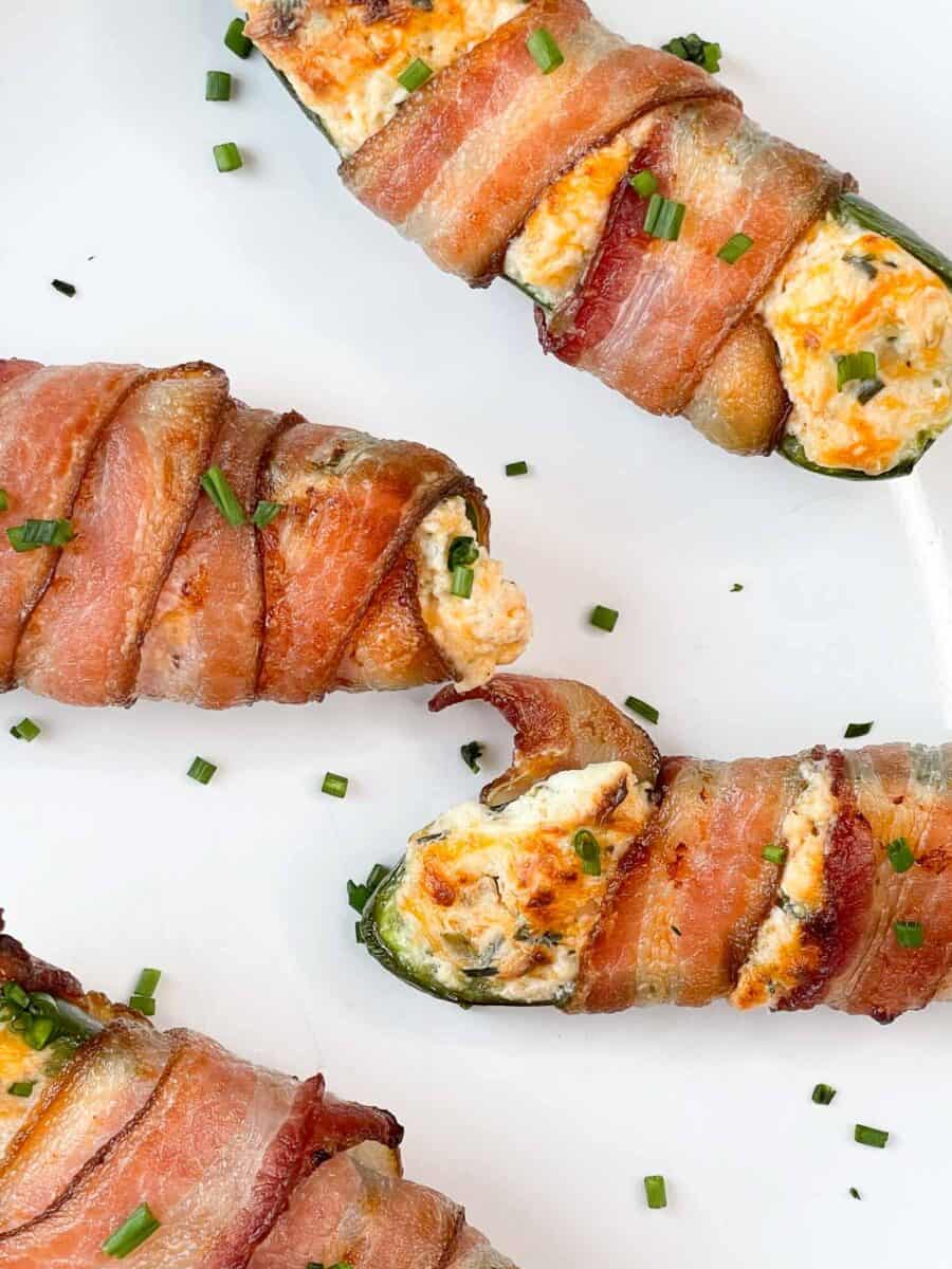 closeup view of four bacon wrapped jalapeños on a white plate with chives sprinkled around