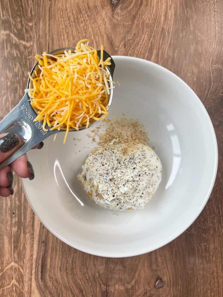 adding cheddar cheese to cheese mixture ingredients in a white bowl
