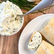 overhead view of garlic herb butter cream cheese spread in small bowl and a plate of bread with some spread on one piece