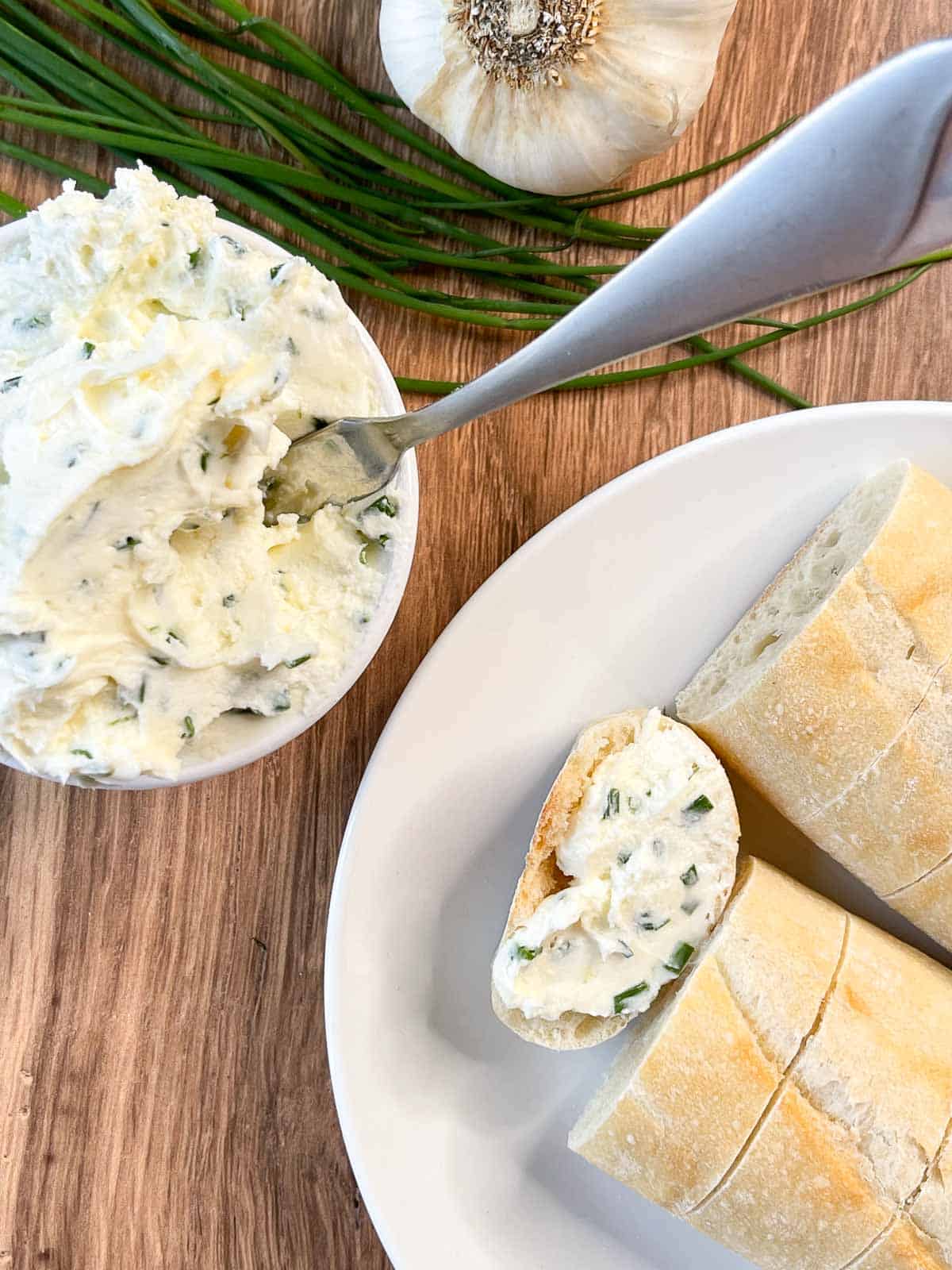 overhead view of garlic herb butter cream cheese spread in small bowl and a plate of bread with some spread on one piece
