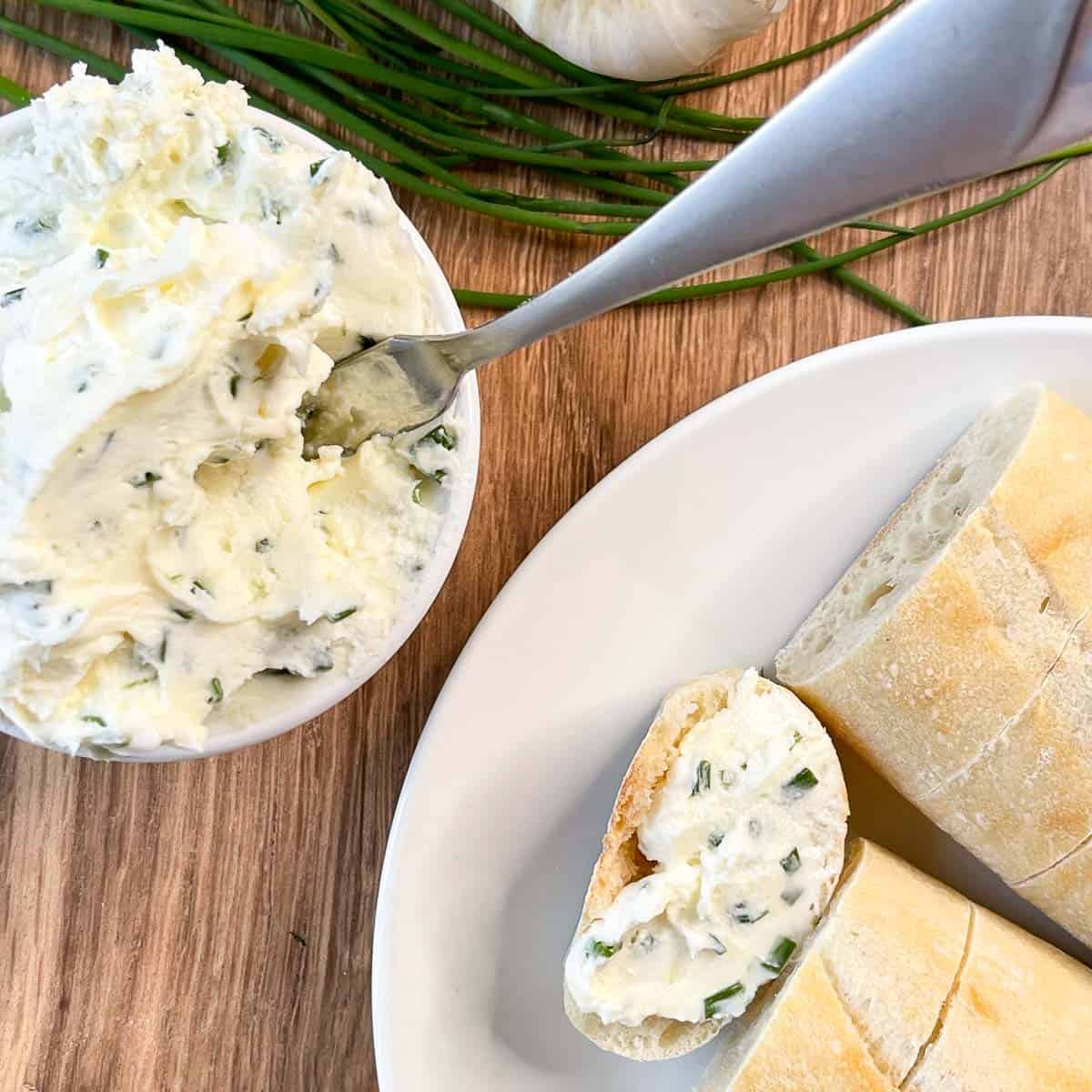 overhead view of garlic herb butter cream cheese spread in small bowl and a plate of bread with some spread on one piece