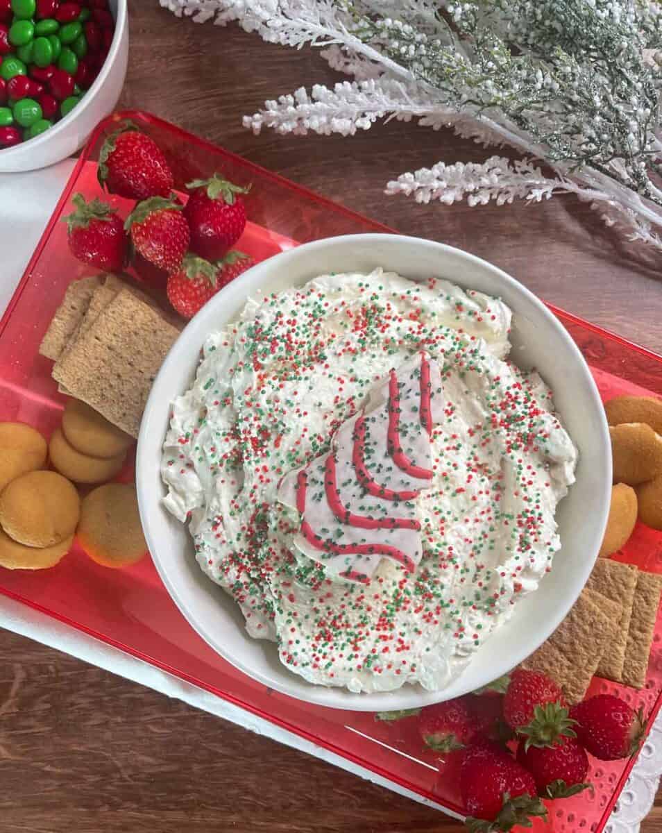 overhead view of little Debbie christmas tree cake dip in a bowl with sprinkles on top and a whole tree cake on top, and a platter with cookies crackers and fruit for dipping